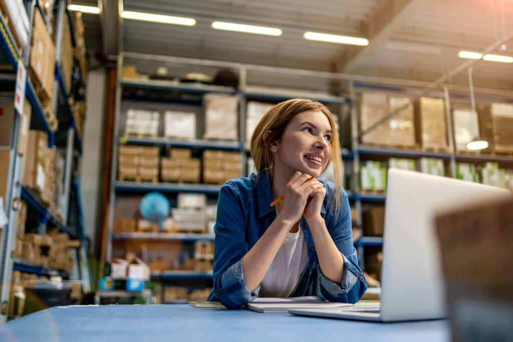 Eine Frau sitzt vor einem Laptop in einem Hochregallager und plant Logistikstrategien. Im Hintergrund sind Regale mit verschiedenen Kartons und Produkten zu sehen, die bis zur Decke reichen