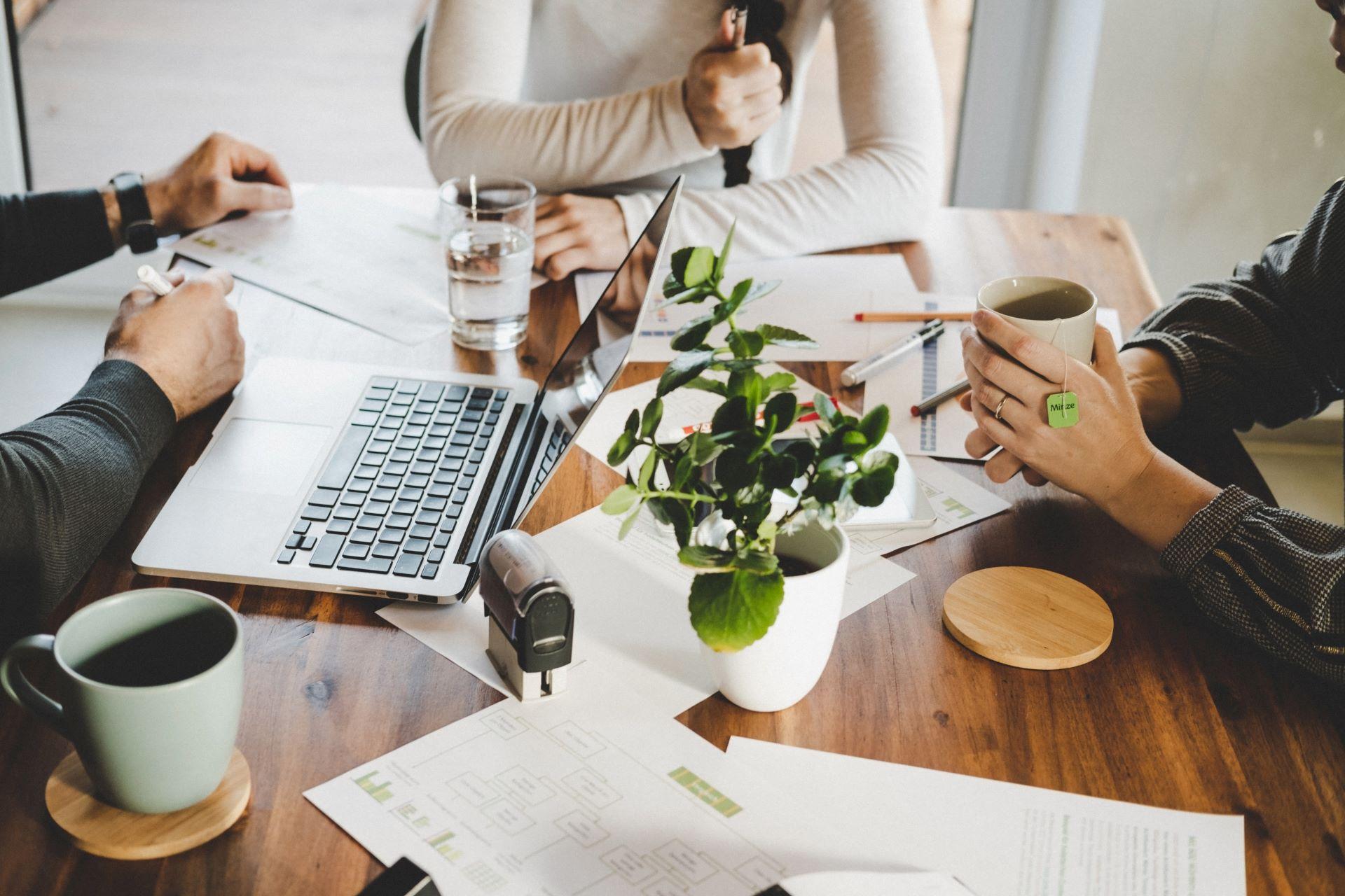 Drei Personen arbeiten an einem Holztisch mit Laptop, Dokumenten, Kaffeetassen und einer Topfpflanze. Eine entspannte Arbeitsatmosphäre, die kreative Zusammenarbeit symbolisiert