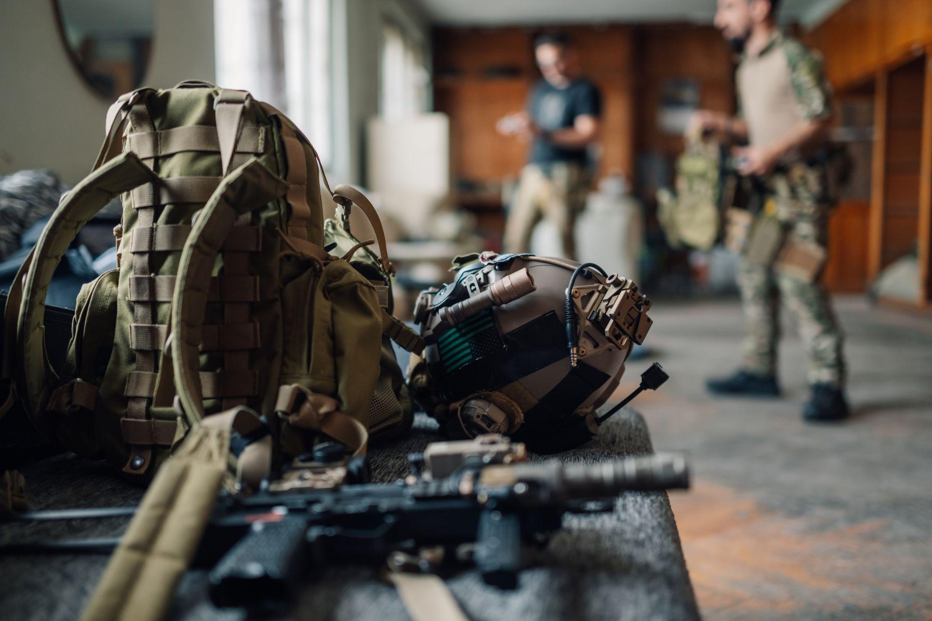 Taktische Airsoft-Ausrüstung mit Helm, Rucksack und Waffe auf Tisch – zwei Spieler im Hintergrund, symbolisieren Einsatzbereitschaft